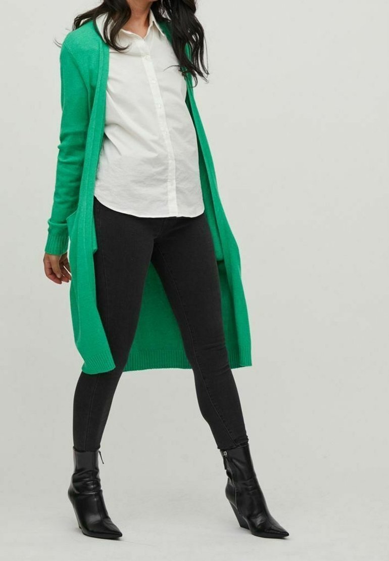 Femme portant un long cardigan vert, une chemise blanche boutonnée, un jean skinny noir et des bottines noires, debout devant un fond uni.