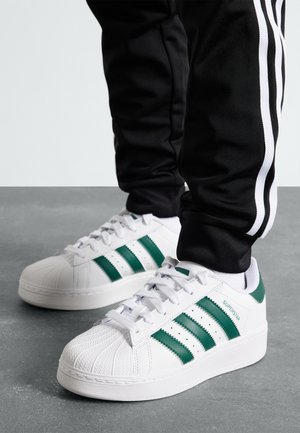 Baskets blanches avec des bandes vertes portées avec un pantalon noir à bandes blanches sur les côtés, posées sur un sol gris.