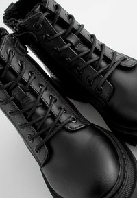 Paire de bottes en cuir noir à lacets avec col rembourré et semelles en caoutchouc robustes sur fond blanc.