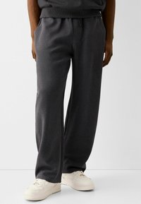 Tmavě šedé tepláky vyrobené z měkké látky, s elastickým pasem, bočními kapsami a volným střihem, doplněné o bílé tenisky.