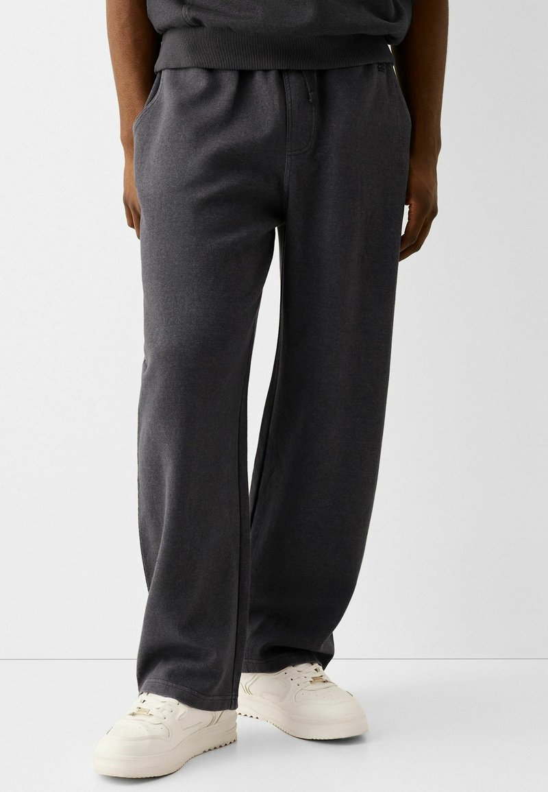 Tmavě šedé tepláky vyrobené z měkké látky, s elastickým pasem, bočními kapsami a volným střihem, doplněné o bílé tenisky.