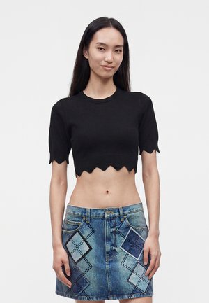 Femme aux longs cheveux foncés portant un crop top noir à bord festonné et une jupe en jean bleue ornée de patchs à losanges écossais, debout devant un fond uni.