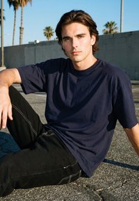 Noormehe tumedate juustega, kes kannab tumesinist T-särki ja musti pükse, istub pragunenud asfaldil, taustal paistavad palmipuud ja müür.