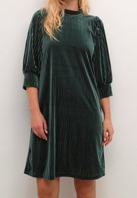 Vestido de terciopelo verde oscuro con rayas verticales, cuello alto, mangas abullonadas de tres cuartos y un corte holgado, que llega hasta la mitad del muslo.
