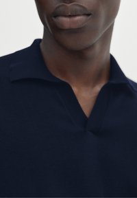 Gros plan sur la partie inférieure du visage et le cou d'une personne portant une chemise à col bleu foncé devant un fond uni.