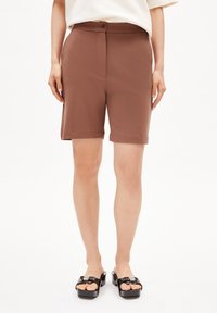 Bruine op maat gemaakte shorts met een knoopsluiting, voorzien van zijzakken en een omgeslagen zoom. Draagt een paar zwarte platform sandalen op een neutrale achtergrond.