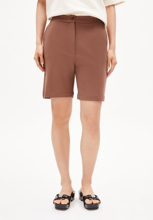 Bruine op maat gemaakte shorts met een knoopsluiting, voorzien van zijzakken en een omgeslagen zoom. Draagt een paar zwarte platform sandalen op een neutrale achtergrond.
