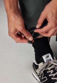 Grå bukser justeres ved kanten, hvilket afslører en sort strømpe og en sort sneaker med grå accenter og hvid sål.