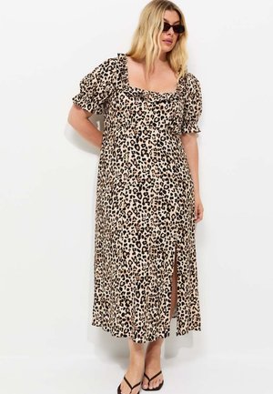 Femme aux cheveux blonds et lunettes de soleil porte une robe midi imprimé léopard avec des manches bouffantes et une fente devant, debout sur un fond blanc.