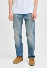Persona que lleva unos vaqueros rectos de color azul claro combinados con unos zapatos de ante marrón claro con cordones y una camisa blanca, de pie sobre un fondo blanco.