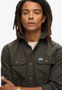 Donkergroen overhemd met knopen, twee borstzakken, bruine knopen en een blauw label op de linkerborstzak. De stof lijkt een textuur te hebben.