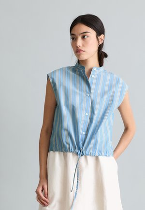 Młoda kobieta o ciemnych włosach, ubrana w bezrękawnik w niebiesko-białe paski zapinany na guziki i zawiązany w talii, w połączeniu z białą spódnicą, na szarym tle.