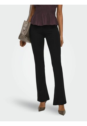 Femeie purtând pantaloni evazați negri, pantofi cu toc ascuțit taupe și un top violet fără mâneci, purtând o geantă taupe pe un umăr.