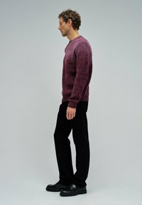 Ein Mann im Profil, der einen strukturierten lila Pullover, schwarze Hosen und schwarze Schuhe vor einem schlichten grauen Hintergrund trägt.