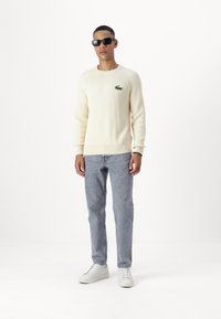 Off-white gerippter Pullover mit grünem gesticktem Logo, hellblaue Jeans und weiße Sneakers. Das Model trägt schwarze Sonnenbrillen.