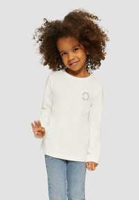 Jong kind met krullend haar, gekleed in een wit T-shirt met lange mouwen en lichte spijkerbroek, lachend en met drie vingers omhoog.