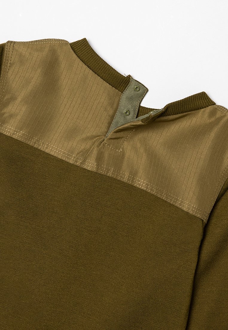 Chemise vert olive avec une épaulière lisse en gabardine beige contrastant. Présente un col côtelé et des fermetures à pression à l'arrière. Texture douce sur l'ensemble.