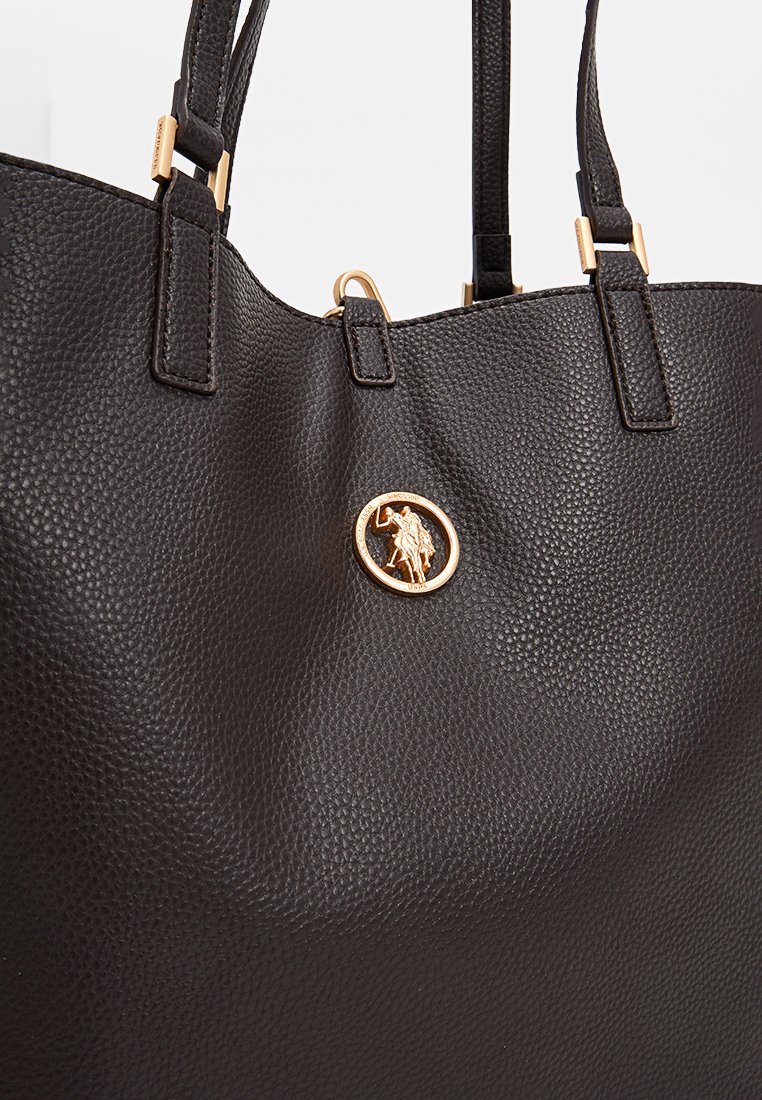 Schwarze, strukturiert aussehende Leder-Tote mit zwei Schulterriemen und einem goldenen, runden Logo-Element auf der Vorderseite. Glattes und klar strukturiertes Design.