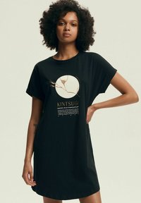 Czarna bawełniana sukienka T-shirt z okrągłą grafiką w złotych i białych akcentach, przedstawiającą tekst i wzór Kintsugi, z krótkimi rękawami.