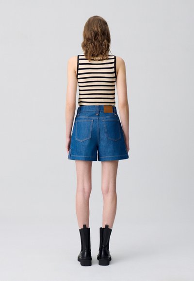 Shorts de mezclilla con un lavado azul, que cuentan con dobles bolsillos traseros y una etiqueta de cuero color marrón claro en la cinturilla. Combinados con una blusa de punto a rayas.
