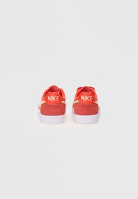 Červené semišové tenisky s bielymi podrážkami, ktoré majú výrazné biele logo Nike na päte a textúrovaný povrch.