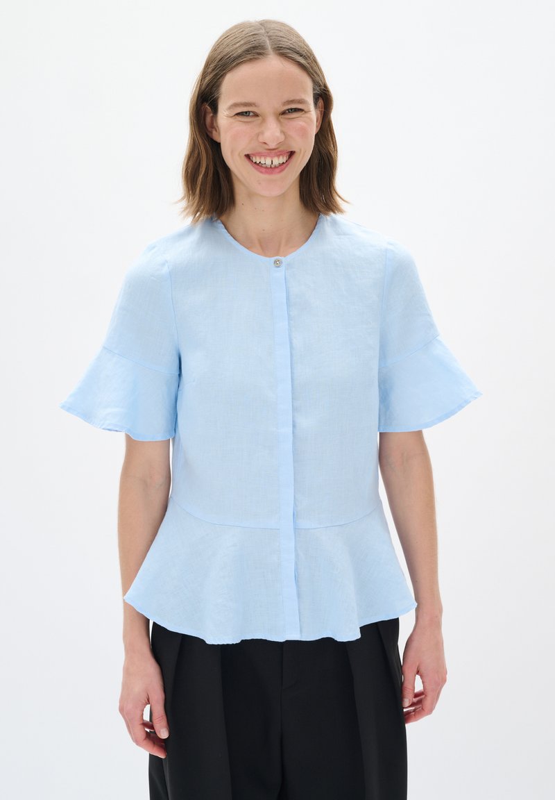 Světle modrá peplum blůza z texturovaného materiálu, s krátkými rozšířenými rukávy a zapínáním na knoflíky vpředu. Hladký povrch, volný střih.