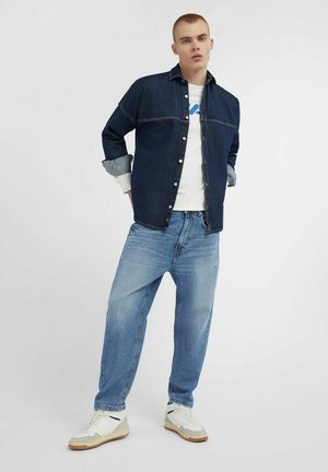 Camicia di jeans di colore blu scuro con chiusura a bottoni, indossata sopra una maglietta bianca. Jeans blu chiaro e sneakers bianche con accenti beige completano il look.