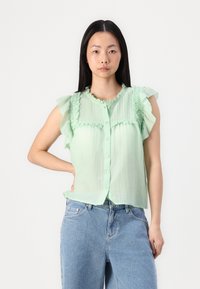 Blusa sem mangas de verdes claros com ombros franzidos, frente com botões e tecido texturizado. Combinada com jeans de ganga azul de cintura alta.