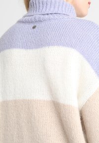 Pull en laine avec un col haut. Présente des rayures horizontales en lavande, blanc et beige. Tissu texturé avec un petit accent métallique à l'arrière.