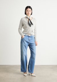Lichtgrijze, textuurgebreide cardigan met een enkele knop, gecombineerd met hooggewaisted blauwe jeans en witte puntige hakken. Zwart-witte sjaal als accent.