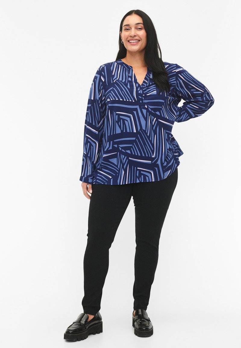 Blusa blu a fantasia con maniche lunghe, caratterizzata da disegni geometrici in diverse tonalità. Abbinata a pantaloni neri slim-fit e scarpe nere lucide.