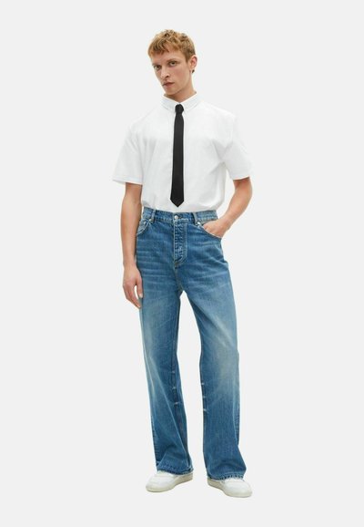 Wit short-sleeve shirt met een kraag en zwarte stropdas, gecombineerd met lichtblauwe wijde jeans en witte sneakers, staande tegen een effen achtergrond.