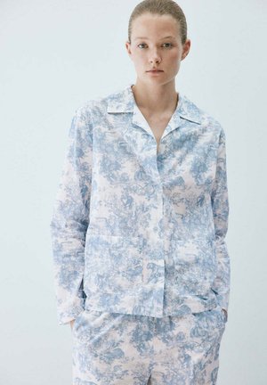 Persona joven con piel clara y cabello recogido lleva una camisa de manga larga con estampado azul y blanco y pantalones a juego con las manos en los bolsillos.