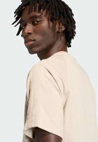 Beżowa koszulka z krótkim rękawem, z subtelnymi teksturowanymi akcentami wzdłuż szwów na ramionach, wykonana z miękkiej bawełnianej tkaniny.