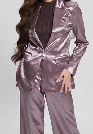 Femme portant un blazer lavande brillant et un pantalon assorti sur un haut côtelé marron foncé, avec de longs cheveux ondulés et des ongles manucurés.