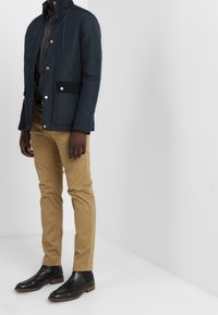 Námořnická prošívaná bunda s vysokým límcem, zlatými knoflíkovými akcenty a předními kapsami, kombinovaná s béžovými chino kalhotami a černými kotníkovými botami.