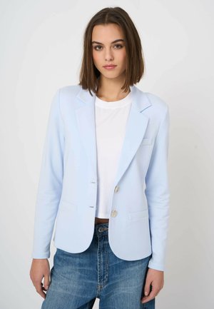Jonge vrouw in een lichtblauwe blazer, wit shirt en blauwe spijkerbroek, staand tegen een effen lichte achtergrond.