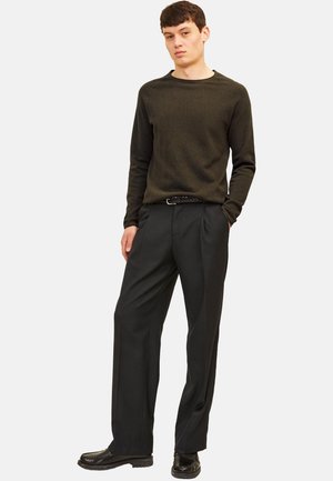 Jonge man staat met handen in zijn zakken, draagt een donker olijfgroene trui, zwarte geplooide broek, gevlochten riem en zwarte nette schoenen.