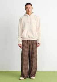 Sweatshirt capuz bege claro com um bolso frontal, combinado com calças largas xadrez em tons de castanho. Ténis em off-white com um design subtil.