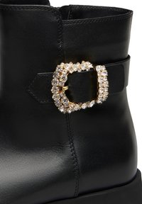 Stivale alla caviglia in pelle nera con una grande fibbia dorata decorata con strass su una cinghia nera, con una texture liscia e una forma strutturata.