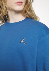 Personne aux cheveux blonds bouclés portant un sweat-shirt bleu avec un petit logo blanc représentant une silhouette sautant sur la poitrine.