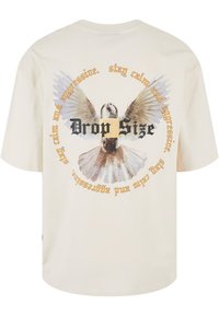 T-shirt en coton beige avec un graphique d'un oiseau aux ailes déployées, entouré de texte en typographie noire et dorée.