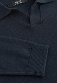 Jūras zila adīta džemperis ar polo apkakli, aprīkots ar ribotām piedurknēm un gludu tekstūru. Etiķetē rakstīts "NEXT, ražots Mjanmā."