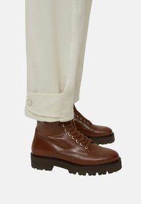 Stivaletti alla caviglia in pelle marrone con punta arrotondata, superficie testurizzata e occhielli tono oro. Abbinati a pantaloni color crema con risvolto.