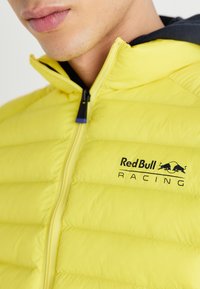 Žuta puffer jakna sa zatvaračem, s crnim logotipom "Red Bull Racing" i dvobojnom teksturom tkanine u blizini kapuljače.