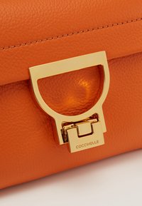 Borsa in pelle di colore arancione con hardware dorato. Presenta una chiusura circolare distintiva e dettagli cuciti lungo i bordi.