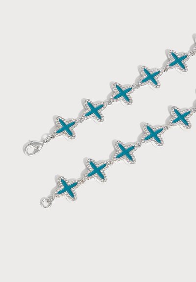 Bracelet en argent avec des maillons bleus en forme de croix bordés de petites pierres transparentes et un fermoir mousqueton sur un fond clair uni.