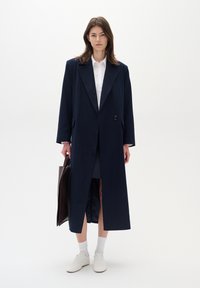 Cappotto lungo navy con un bottone singolo, due tasche frontali e un design elegante, abbinato a una camicia bianca, una gonna grigia e sneakers bianche.