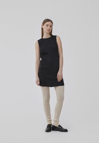 Crna haljina bez rukava s teksturiranim prugama, uparena s kremastim leggingama s uzorkom i crnim loafers cipelama. Minimalistički dizajn, lagani materijal.