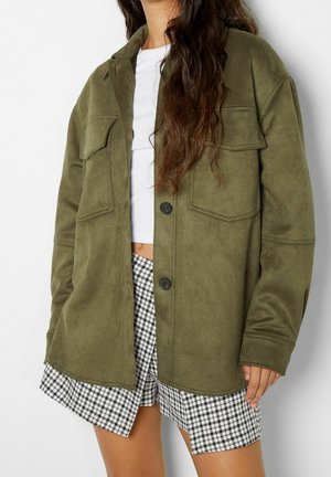 Persona que lleva una chaqueta verde oliva con botones y bolsillos con solapa, top corto blanco y falda de pata de gallo en blanco y negro.